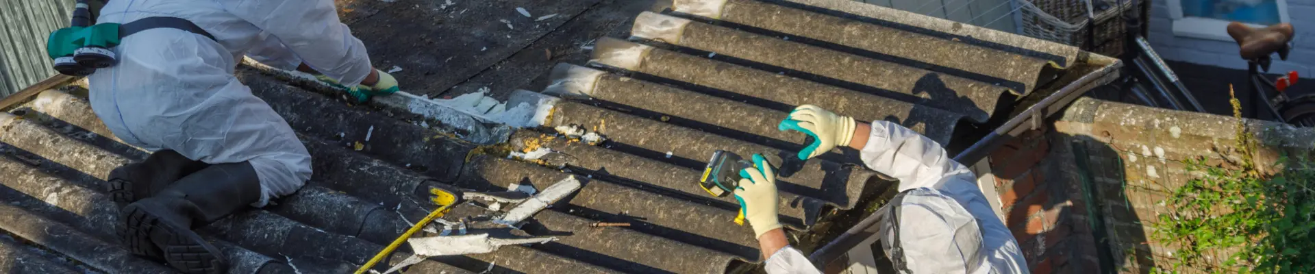 Двама строителни работника облечени ж предпазно облекло и с противогази, премахват азбестови плоскости от покрив